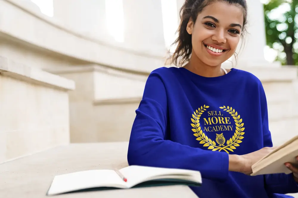Student in a royal blue Sell More Academy sweatshirt studying with a notebook on marble steps